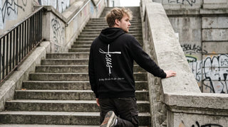 Junger Mann von hinten mit christlichem Hoodie mit Rückenprint auf einer Betontreppe