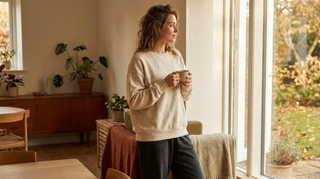 Person in schlichtem Sweatshirt mit Kaffeetasse am Fenster
